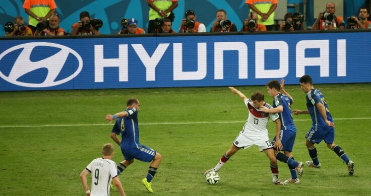 A World Cup game in play in a stadium with 2 players in white and 3 players in blue engaging in a tackle with a giant blue “Hyundai” banner behind.