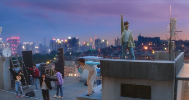 All 7 members of BTS singing and dancing on a rooftop in the middle of a city at dusk.
