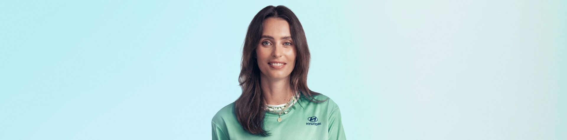 A close up of Ella Mills’ face. She is standing in front of a blue and green background and is wearing her green Team Century jersey and a couple of necklaces. She has long brown hair and blue eyes.