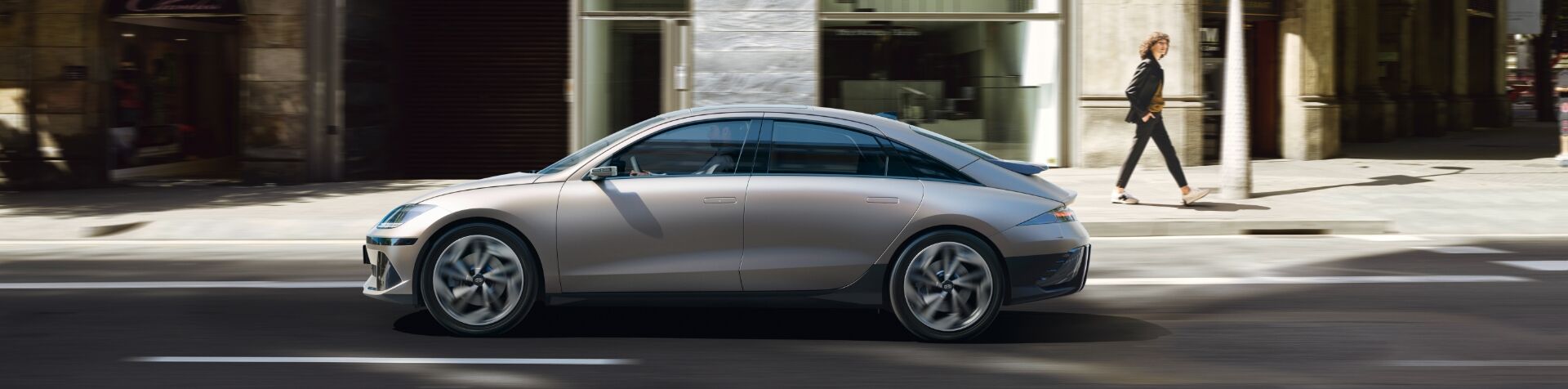 A side view of a silver IONIQ 6 driving along a road in the city. Behind the car a woman is walking along the sidewalk.