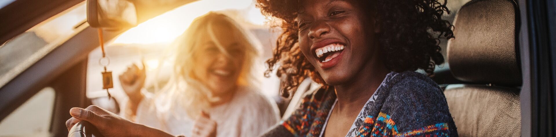 A photographic image of two women smiling in the front seats of a car. The passenger is white and has long blond hair and is wearing a white blouse, the driver is black and has long dark hair.