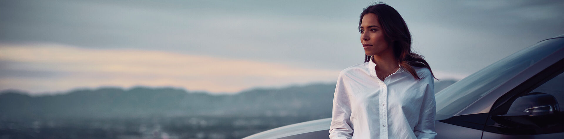 Woman with a white shirt standing in front of a Hyundai NEXO with hills in the background.