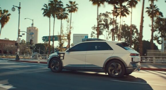 A white Hyundai IONIQ 5-based robotaxi driving along a road lined with palm trees in LA.